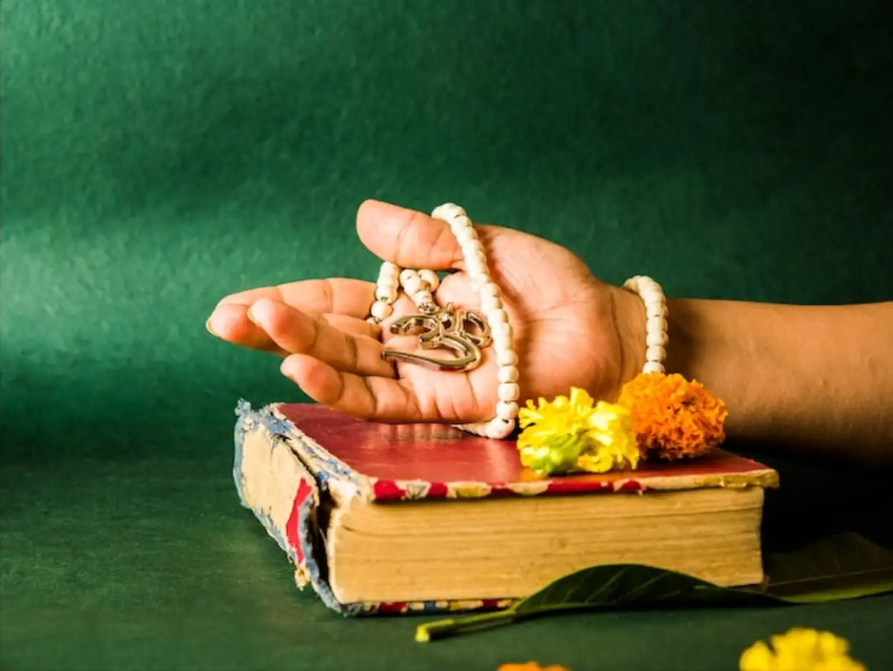 Open hand showing Aum locket with beads, red hindu book in the surface with flowers