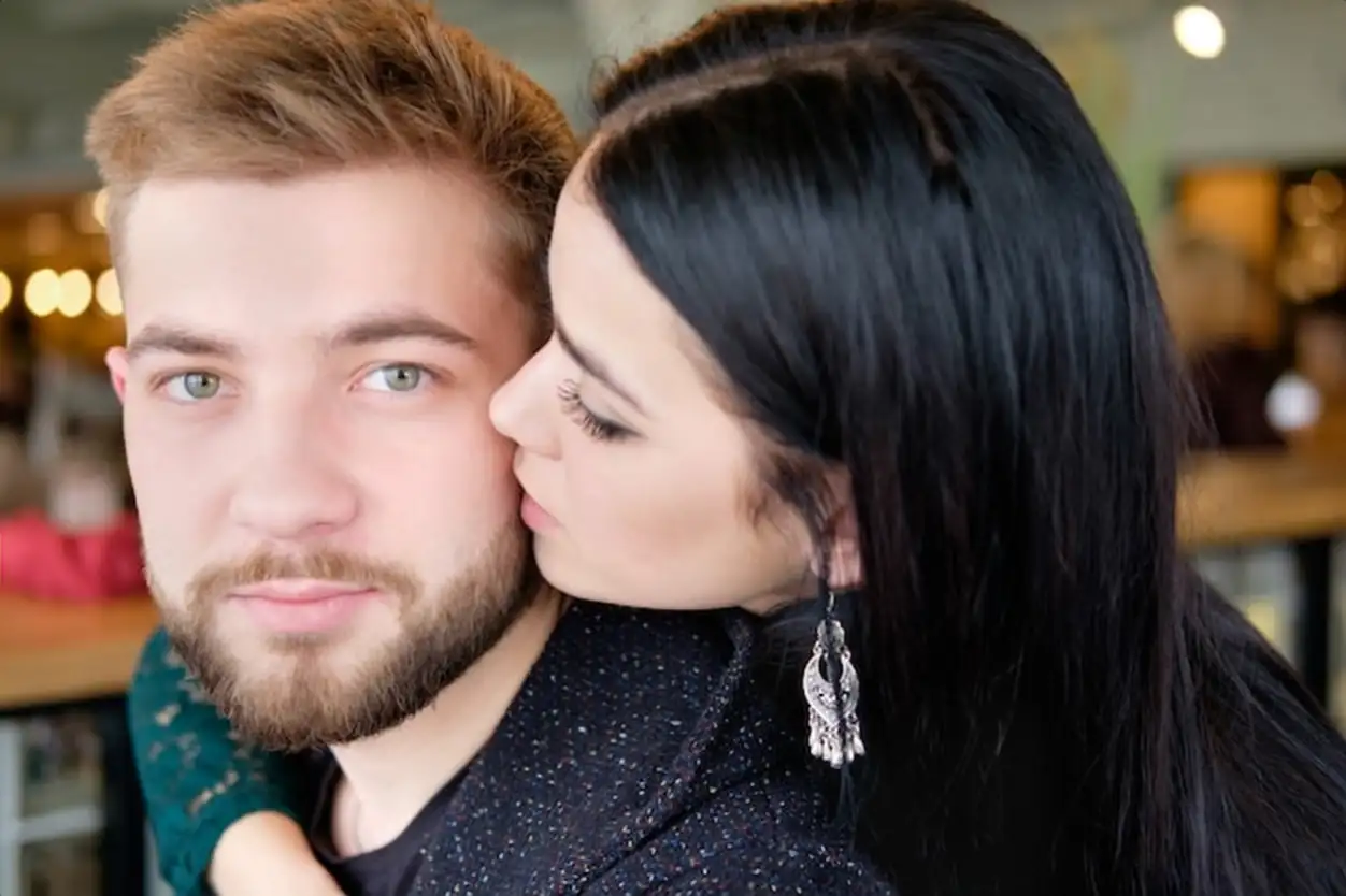 Brunette woman hugs her blond boyfriend around his neck and kiss him
