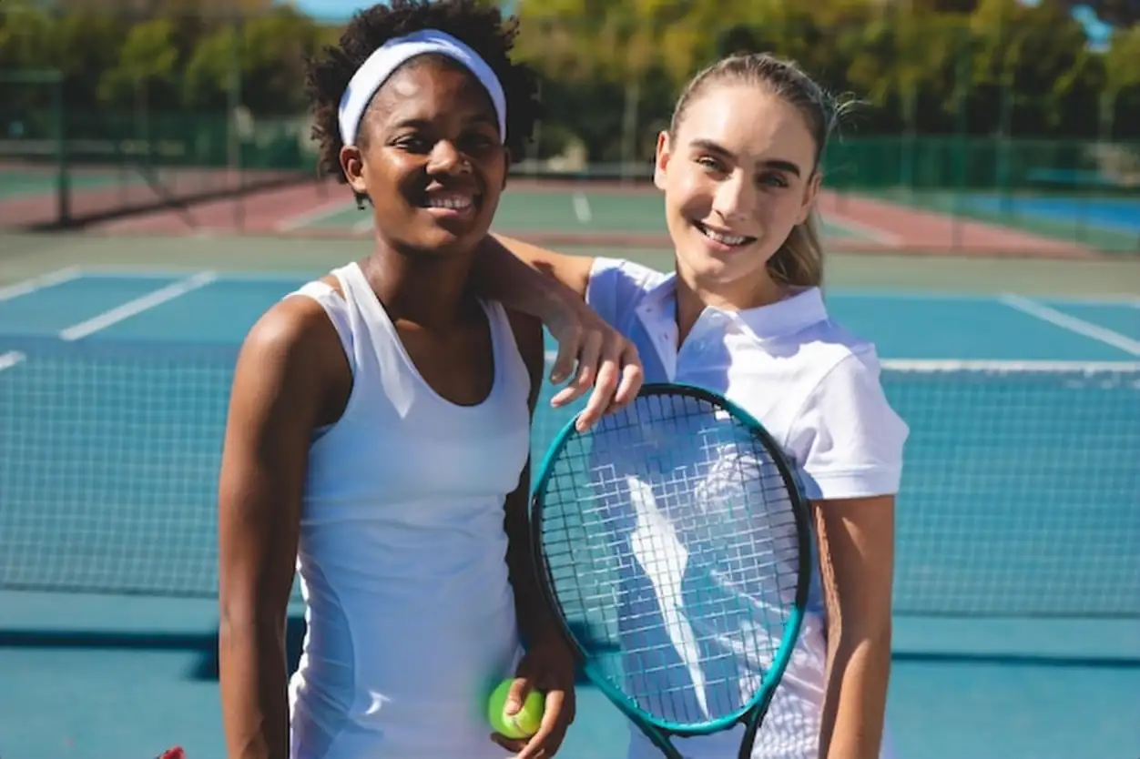 Portrait of smiling multiracial female athletes with racket and ball standing at tennis court