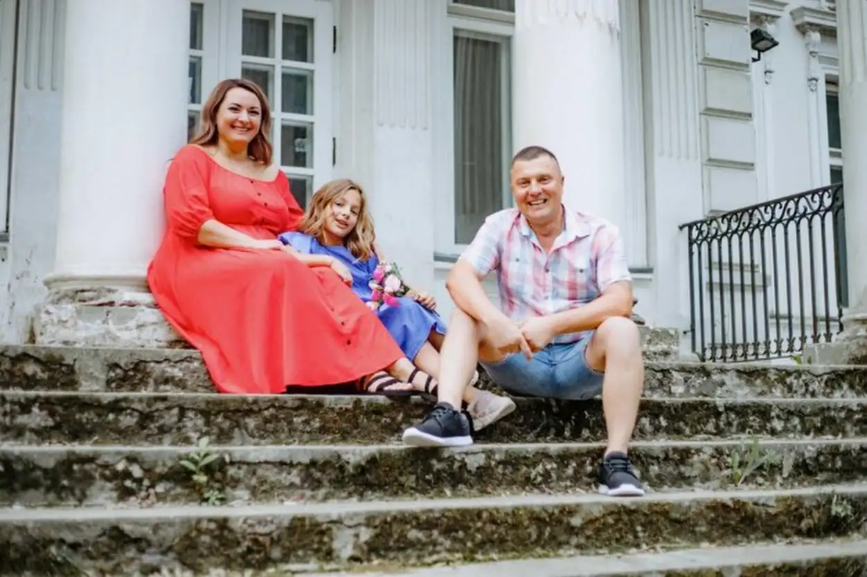 Beautiful woman is sitting on steps of wide staircase next to her charming daughter and husband