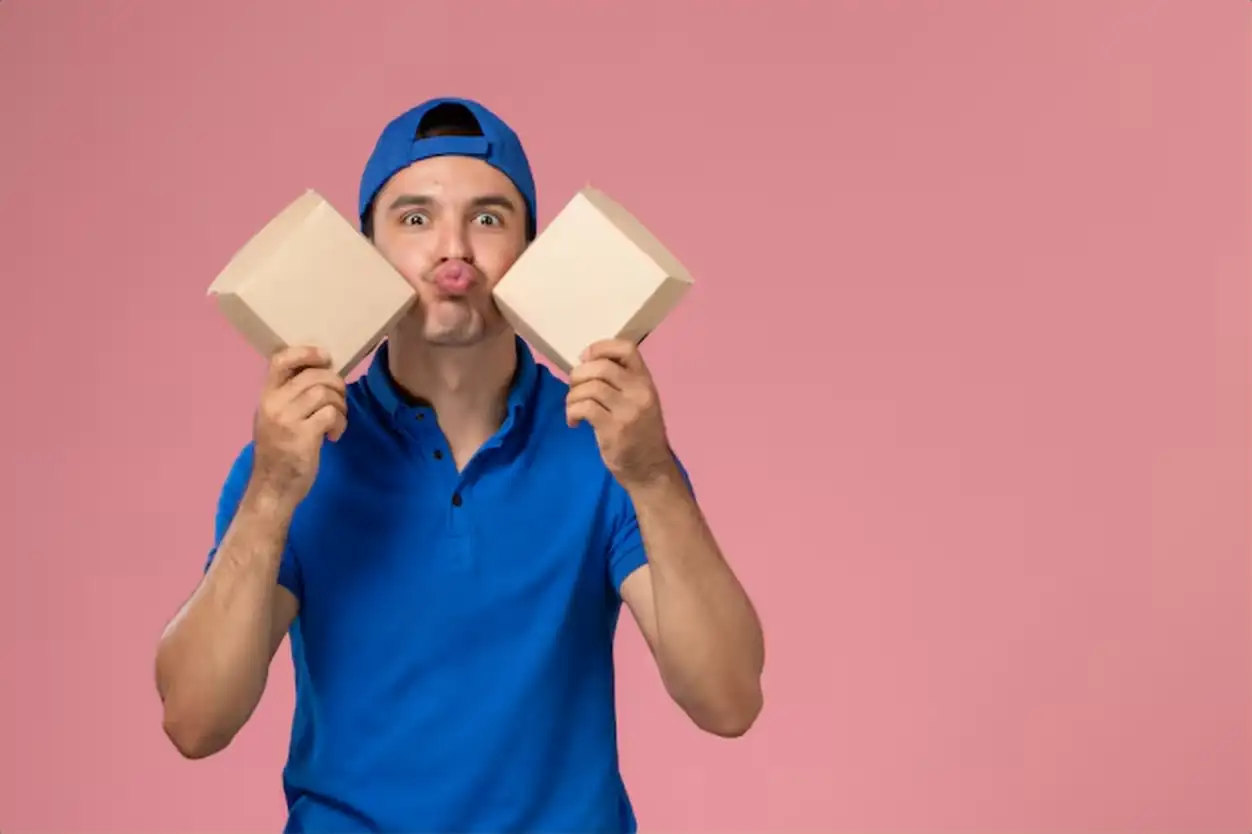 Front view young male courier in blue uniform cape holding little delivery food packages on light pink wall