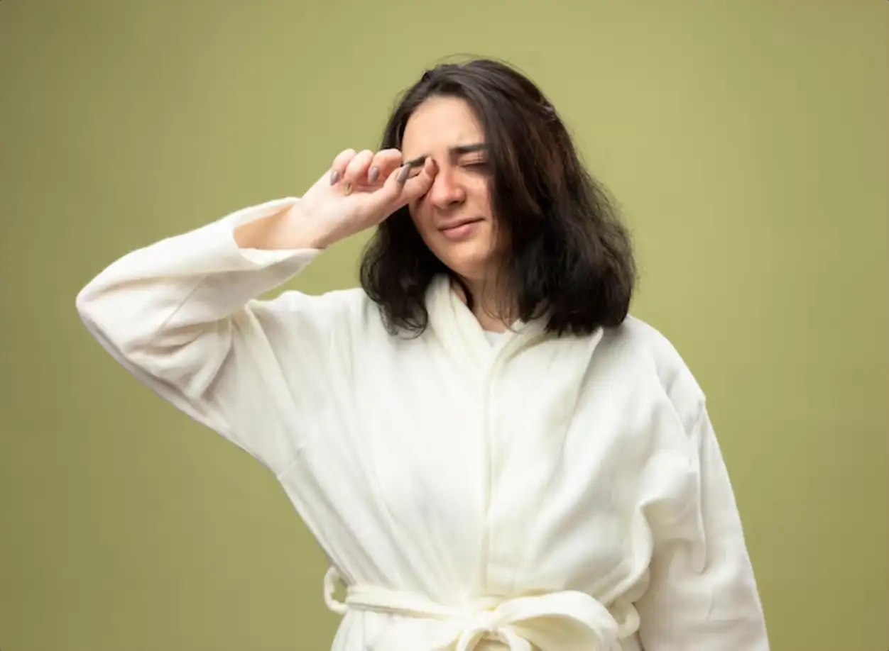 Weak young caucasian ill girl wearing robe wiping eye with closed eyes isolated on olive green background