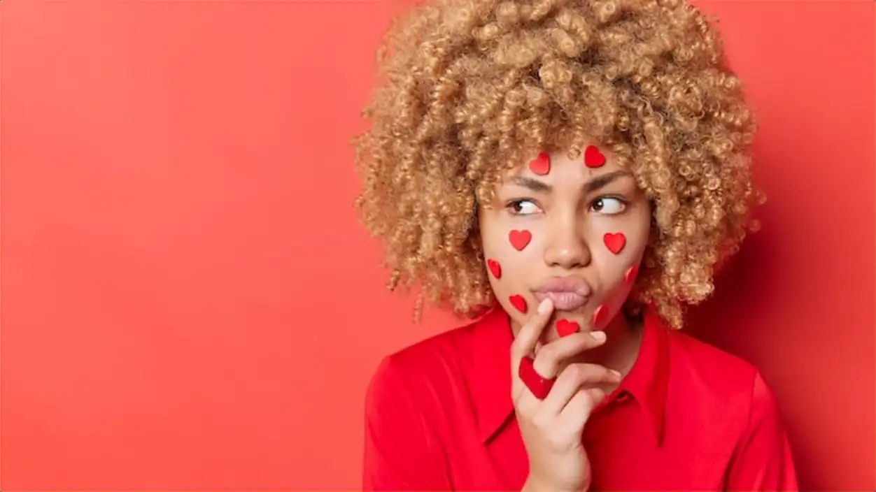Horizontal shot of sullen discontent woman with curly bushy hair purses lips looks sadly away has little hearts stuck on face wears shirt isolated over red background blank space for your promo